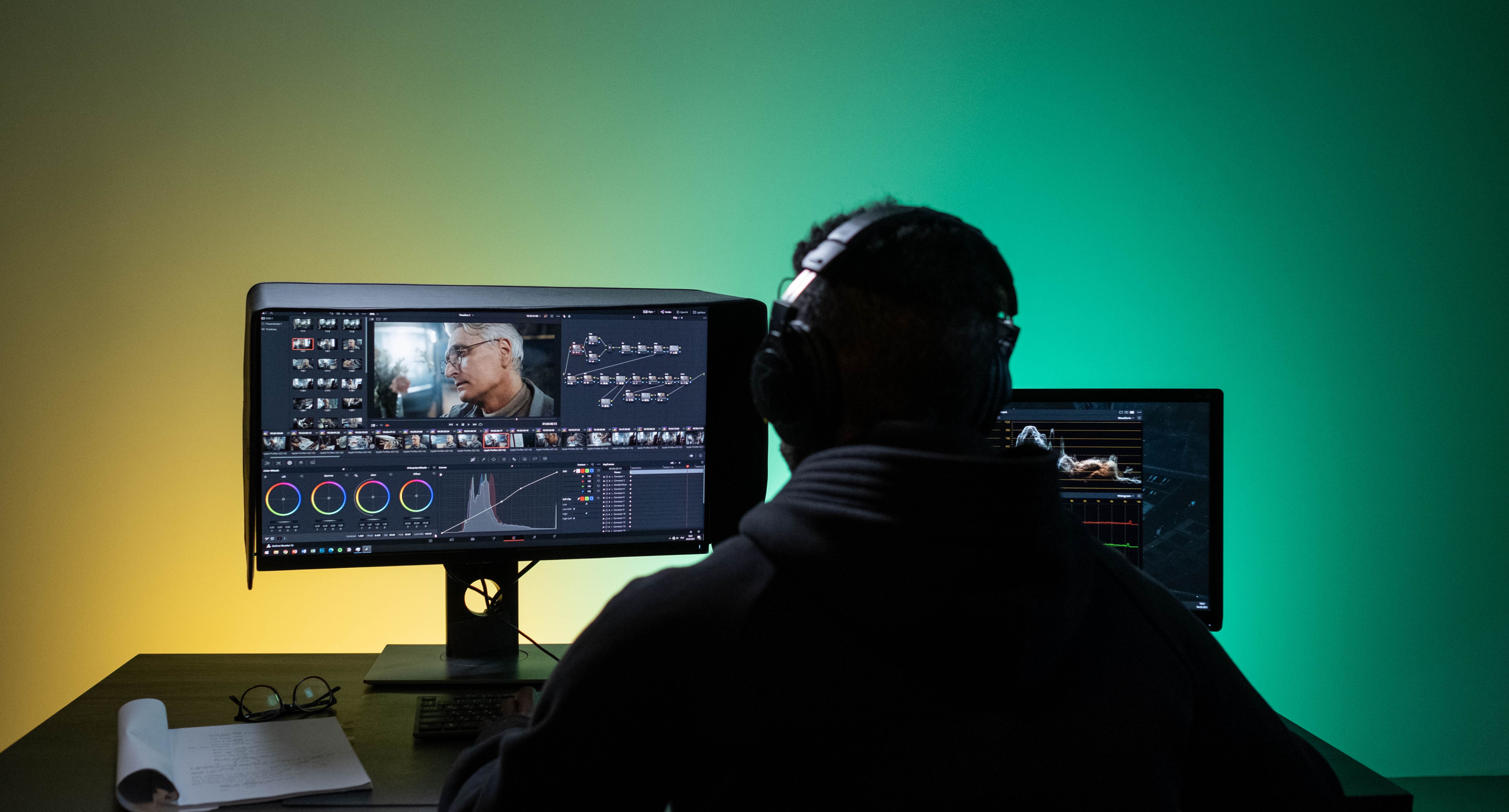 person with headphones sitting with their back facing the camera in front of two computer screens, one screen is a video editing app 